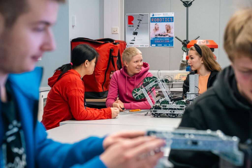 Lukio-oppilaita luokkahuoneessa pöydän ääressä. Pöydällä on myös robotteja.