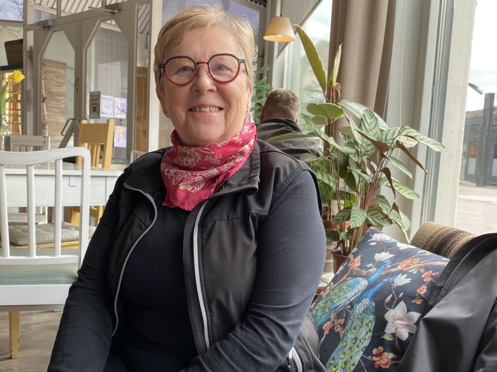 Hymyilevä nainen istuu. Hänellä on silmälasit ja huivi kaulassa. Taustalla on viherkasvi.