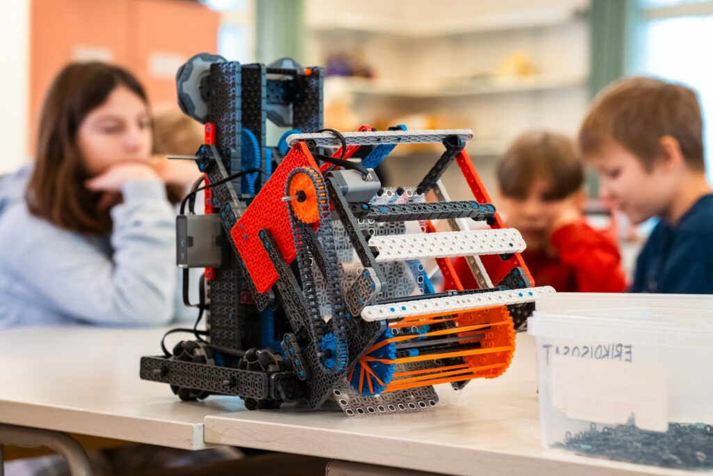 VEX-robotti pöydällä. Se muistuttaa hieman puimuria. Robotin taustalla on koululaisia.