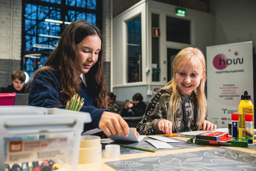 Kaksi tyttöä pöydän ääressä. Pöydällä on askartelutarvikkeita.