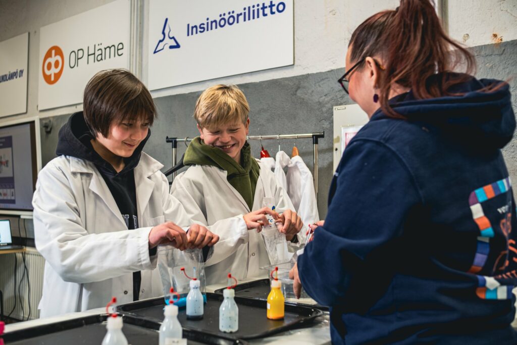 Kaksi lasta ja aikuinen pöydän ääressä. Pöydällä on väripulloja lasilla on käsissään pienet muovipussit.