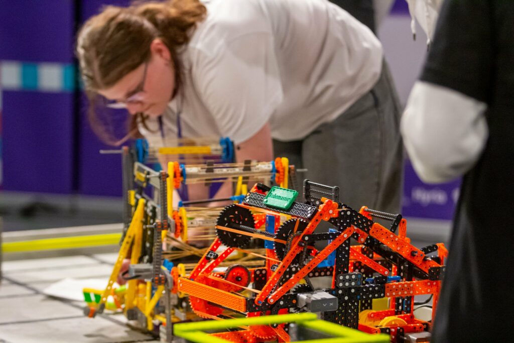 Kaksi VEX-robottia kisakentällä. Niiden takana on kumartuneena nuori tyttö.