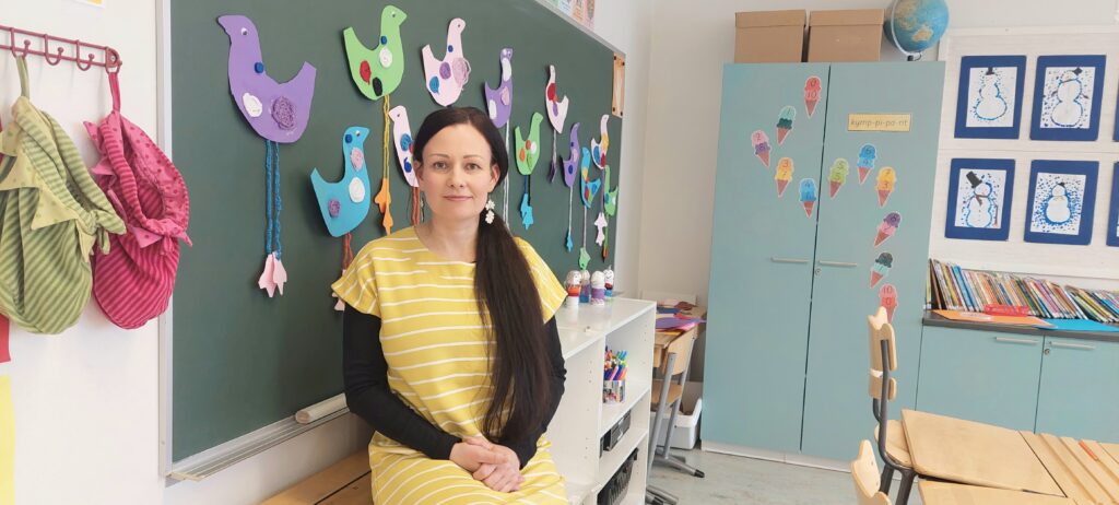 Pitkähiuksinen nainen istuu pöydän reunalla. Taustalla on liitutaulu, johon on kiinnitetty lasten askarteluja.