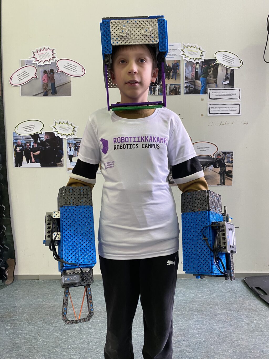 Poika, jolla on VEX-osista tehty kypärä päässään ja käsissään robottikädet.