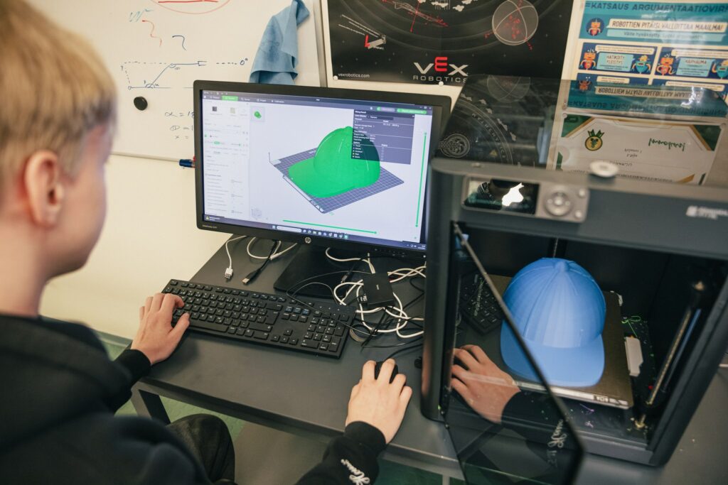 Koululainen katsoo näytöltä lippalakkia, joka on vieressä 3D-tulostetimessa.